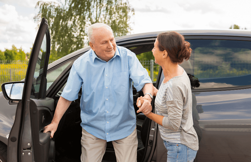 An elderly man is assisted by a woman as he steps out of a car. They are both smiling and appear to be engaged in a warm conversation, suggesting support and companionship during transport or rehabilitation care.