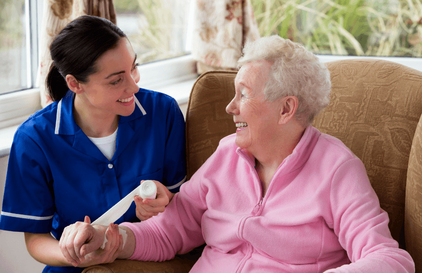 A smiling nurse in a blue uniform gently wraps a bandage around the wrist of an elderly woman in a pink fleece jacket. They are seated indoors, engaging warmly and making eye contact, conveying a sense of trust and compassionate care.