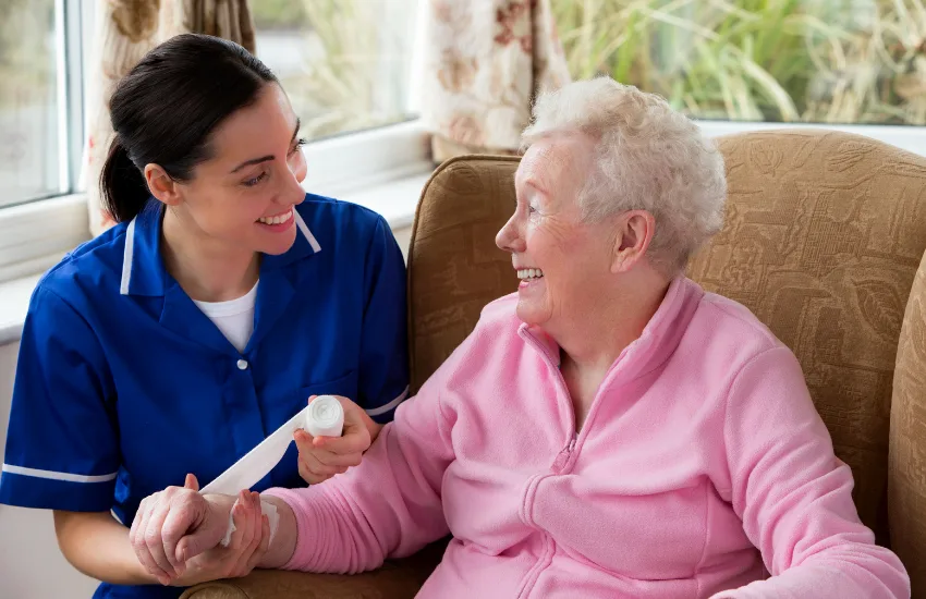 A smiling nurse in a blue uniform gently wraps a bandage around the wrist of an elderly woman in a pink fleece jacket. They are seated indoors, engaging warmly and making eye contact, conveying a sense of trust and compassionate care.