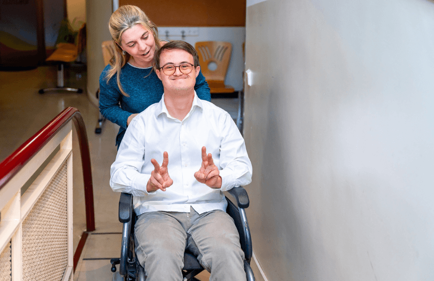 A smiling young man with Down syndrome, seated in a wheelchair, flashes peace signs with both hands while a supportive woman, standing behind him, gently holds the wheelchair handles.