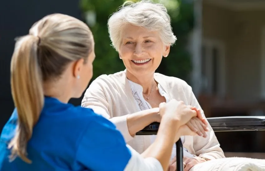 Female nurse, holding hands with older woman smiling while seated in wheelchair, in relation to our discussion on private nursing giving older Australians' more independence.