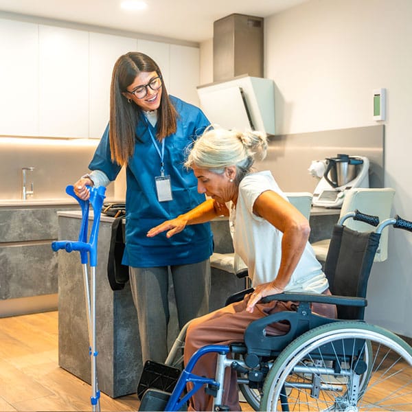 An elderly woman in a wheelchair receives assistance from a female caregiver.