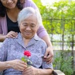 Older woman in chair smiling at a rose while younger woman embraces her from behind.