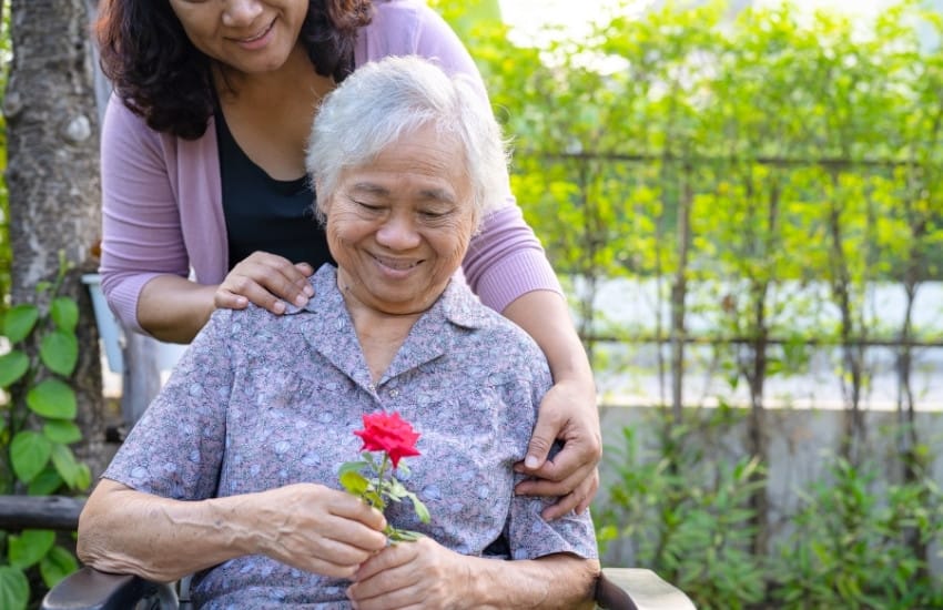 Older woman in chair smiling at a rose while younger woman embraces her from behind.